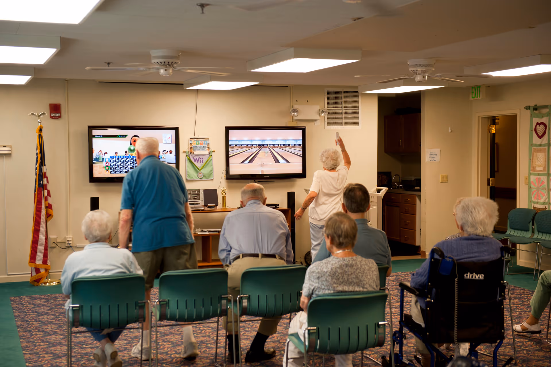 A group of elderly people in a senior living facility room, some seated and one standing, playing a virtual bowling game displayed on two wall-mounted screens. The room has green chairs, an American flag, and a small kitchenette area in the background.