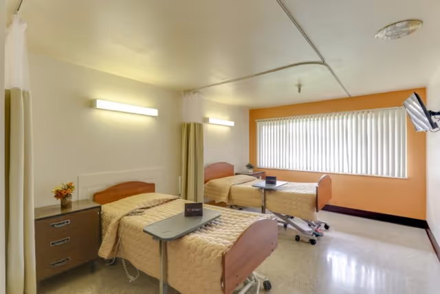 A bright shared care facility bedroom with two adjustable beds, overbed tables, a dresser, vertical blinds and a wall-mounted TV.