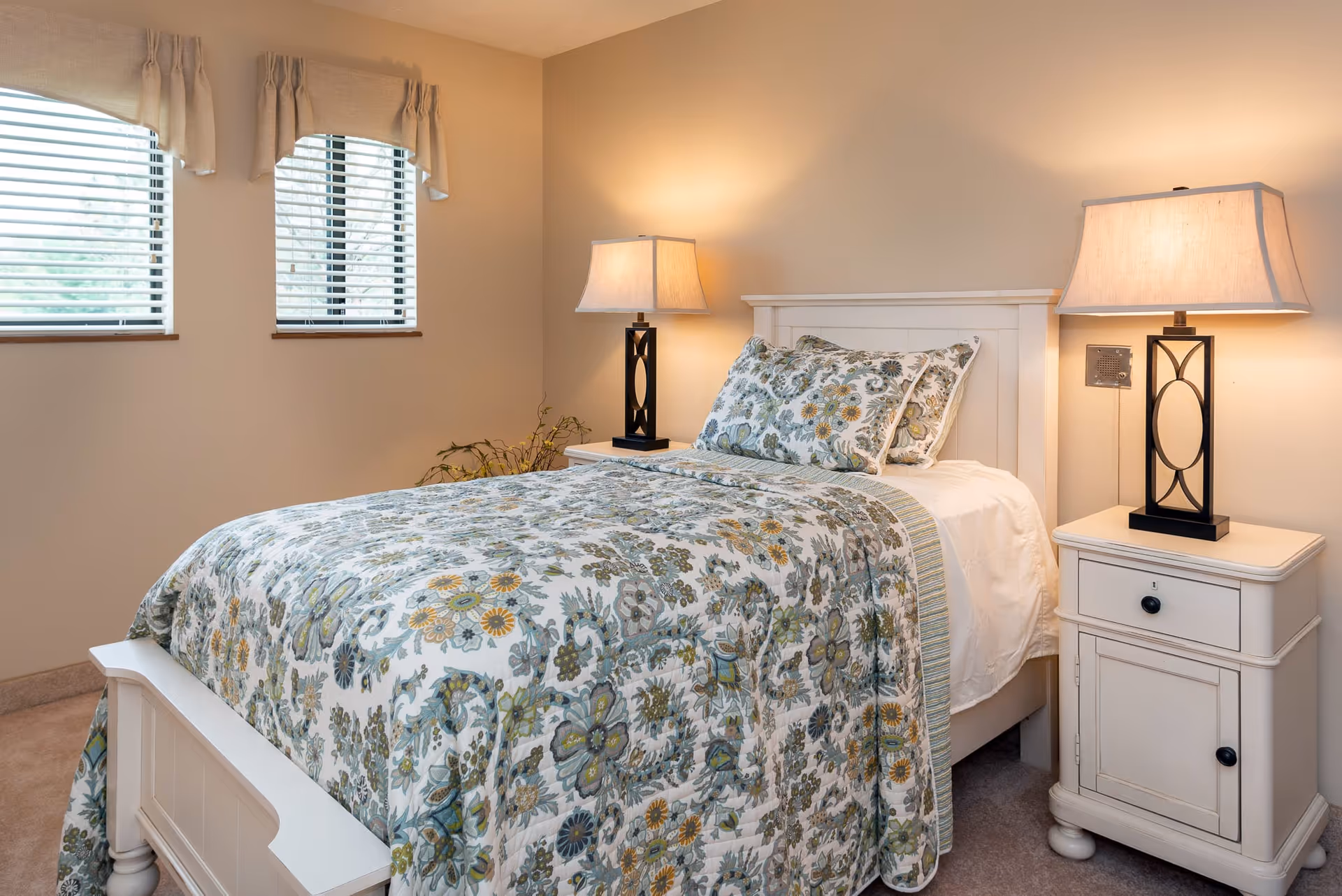 A cozy bedroom with a white wooden bed frame and matching nightstands on each side. The bed is made with a floral patterned quilt and pillows in shades of green, blue, and yellow. Two table lamps with rectangular beige shades sit on the nightstands, casting a warm light. There are two windows with white blinds and beige valances on the left wall.