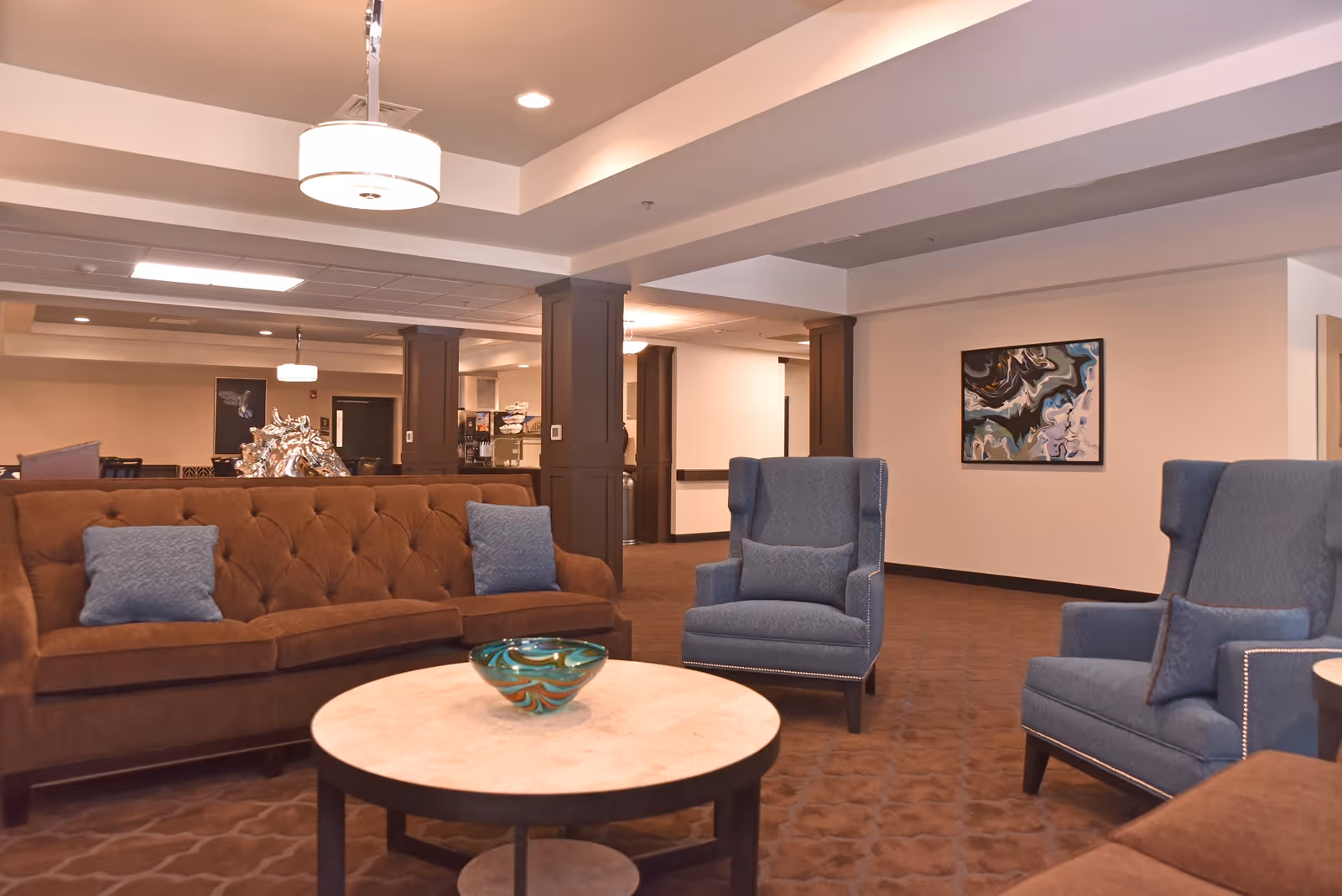 A cozy living room area in The Glen facility featuring a brown tufted sofa with blue cushions, two blue armchairs with matching pillows, a round coffee table with a decorative glass bowl, and abstract artwork on the wall. The room has warm lighting and a carpeted floor.