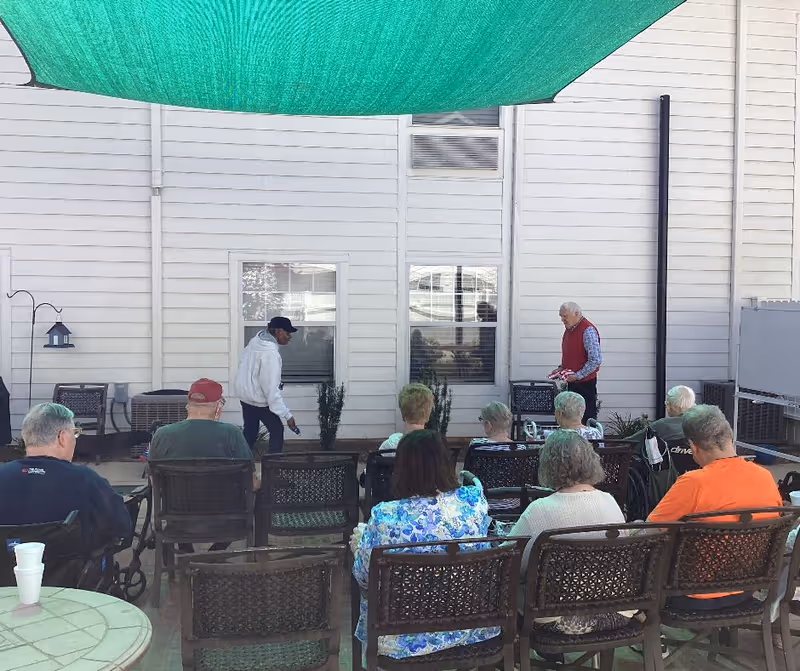 A group of elderly people seated outdoors on chairs facing a white building wall with windows. Two men stand near the wall, one in a white hoodie and black cap, the other in a red vest and checkered shirt. A green shade cloth is overhead providing shade.