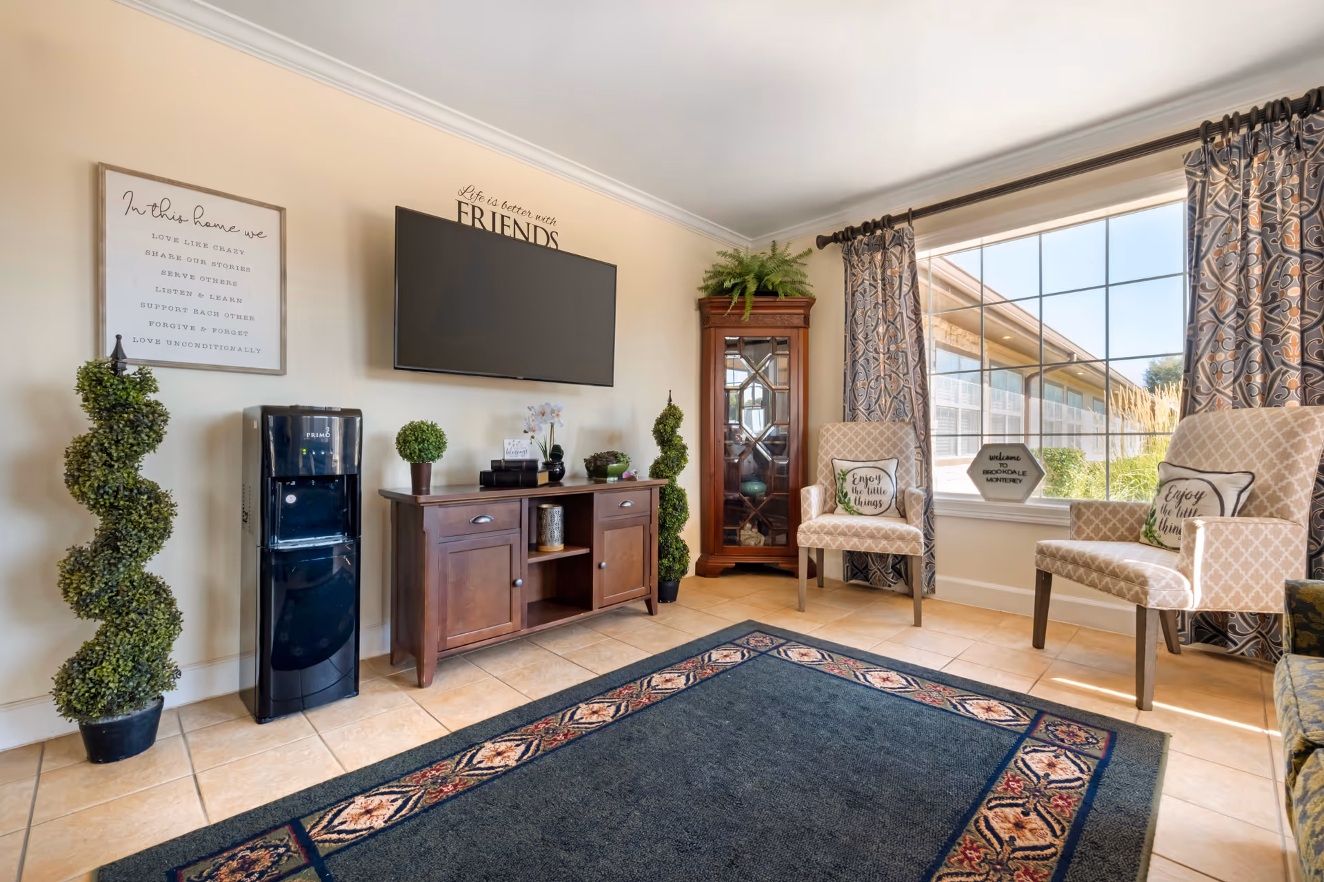 A bright and cozy living room area with a large window letting in natural light. The room features two patterned armchairs with decorative pillows, a wooden cabinet with plants and decor items, a flat-screen TV mounted on the wall, a water dispenser, and a tall glass-front cabinet in the corner. The floor is tiled and there is a large area rug with a decorative border. Wall decor includes a framed inspirational quote and a sign above the TV that reads 'Life is better with FRIENDS'.