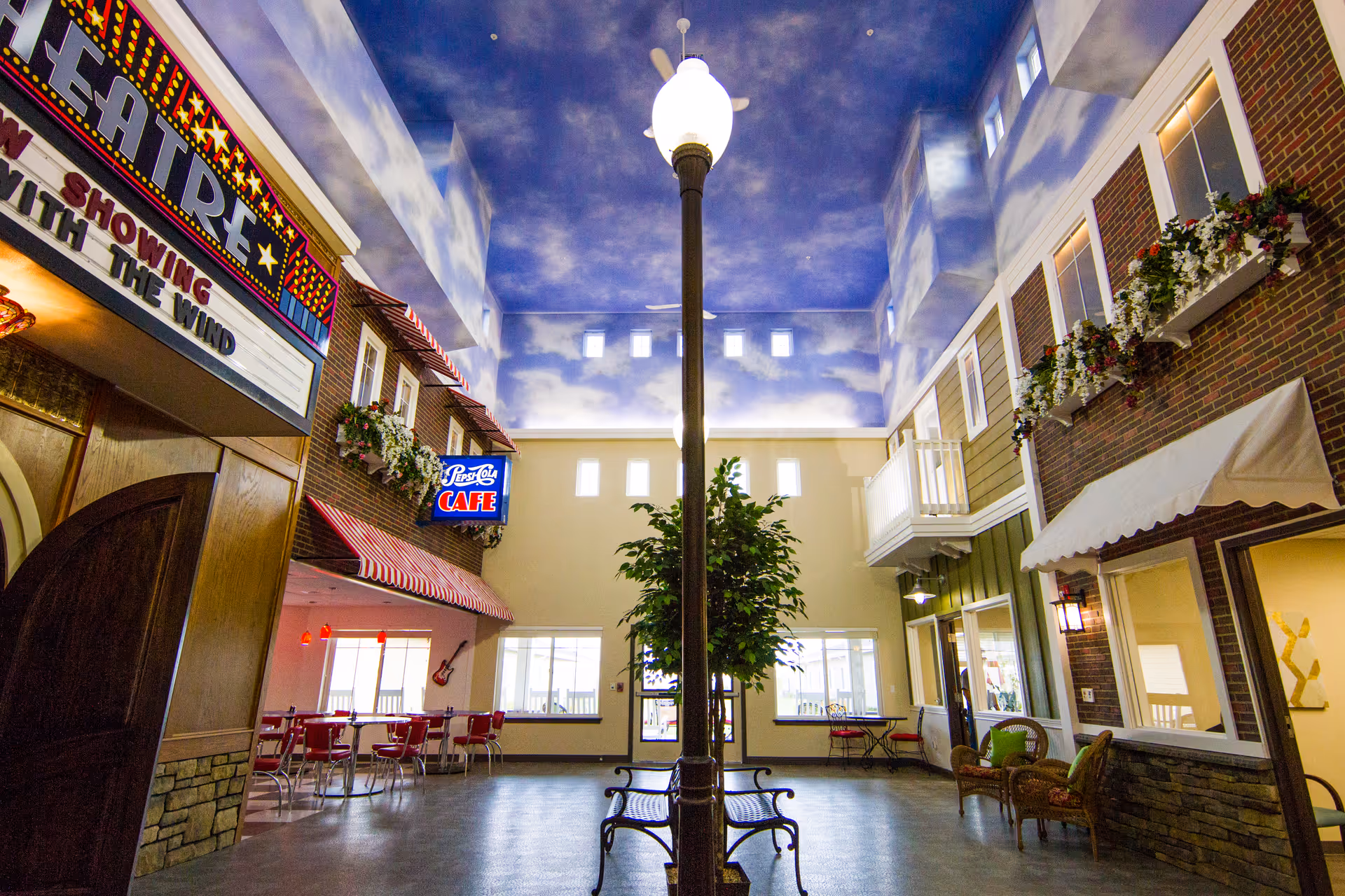 Indoor common area designed to resemble a small town street with a painted sky ceiling, a tall street lamp in the center, benches around the lamp, a theater marquee on the left, a Pepsi-Cola cafe sign with red chairs and tables underneath, and building facades with windows and flower boxes on the right.