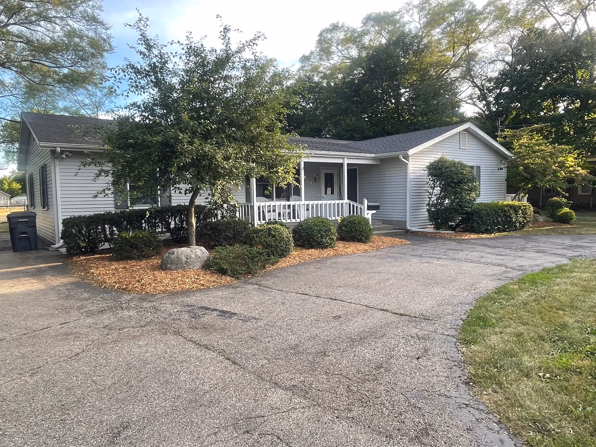 Single-story light-gray house with a small front porch, driveway, and landscaped shrubs and trees.
