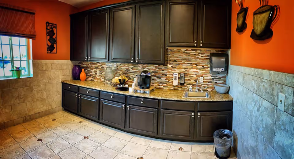 A kitchen area with dark brown cabinets and a granite countertop. The backsplash features a mosaic tile design in earth tones. On the counter, there is a coffee maker, a glass container, and decorative items. The walls are painted orange above beige tiled wainscoting. There is a window with a brown blind on the left side and two decorative wall hangings on the right. The floor is tiled with large beige tiles with small brown diamond accents. A trash can is placed on the floor to the right.