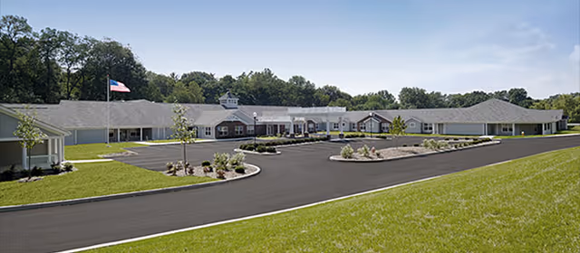 Single-story senior living facility building with a circular driveway, landscaped islands, and an American flag under a clear sky.
