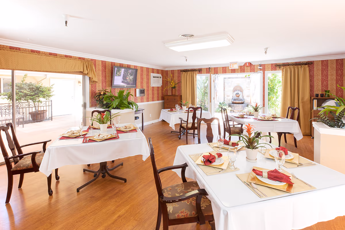 A bright dining room with several tables covered in white tablecloths, each set with plates, glasses, gold placemats, and red napkins. The room has wooden chairs with floral cushions, red and gold striped wallpaper, and large windows with yellow curtains letting in natural light. There are plants on the tables and around the room, and a TV mounted on the wall. Outside the windows, greenery and a water fountain are visible.