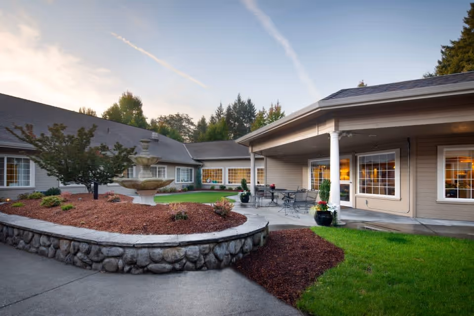 Outdoor courtyard area of a senior living facility with a stone-bordered garden bed featuring a tiered fountain, surrounded by a paved walkway and green lawn. The building has beige siding, white trim, large windows, and a covered patio with metal tables and chairs. Trees and a partly cloudy sky are visible in the background.