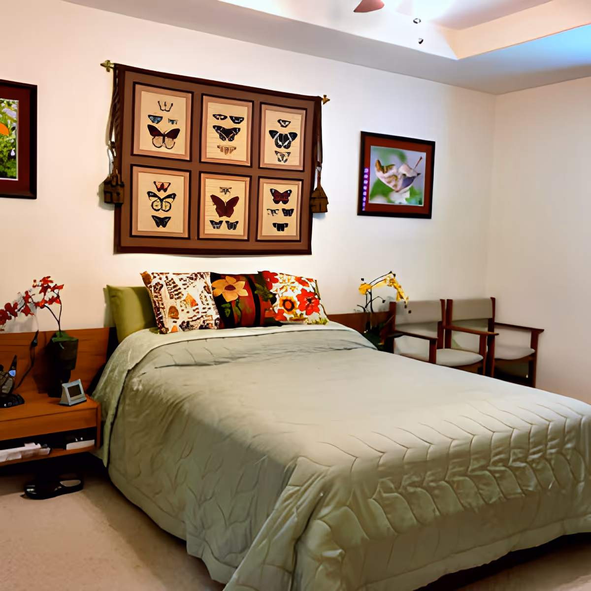 A cozy bedroom with a neatly made bed covered in a light green quilt. The bed has three decorative pillows with floral and butterfly designs. Above the bed hangs a framed textile artwork featuring various butterflies. To the right, there are two wooden chairs with light cushions and a framed picture of a bird on the wall. A small wooden nightstand with a plant and a digital clock is on the left side of the bed.