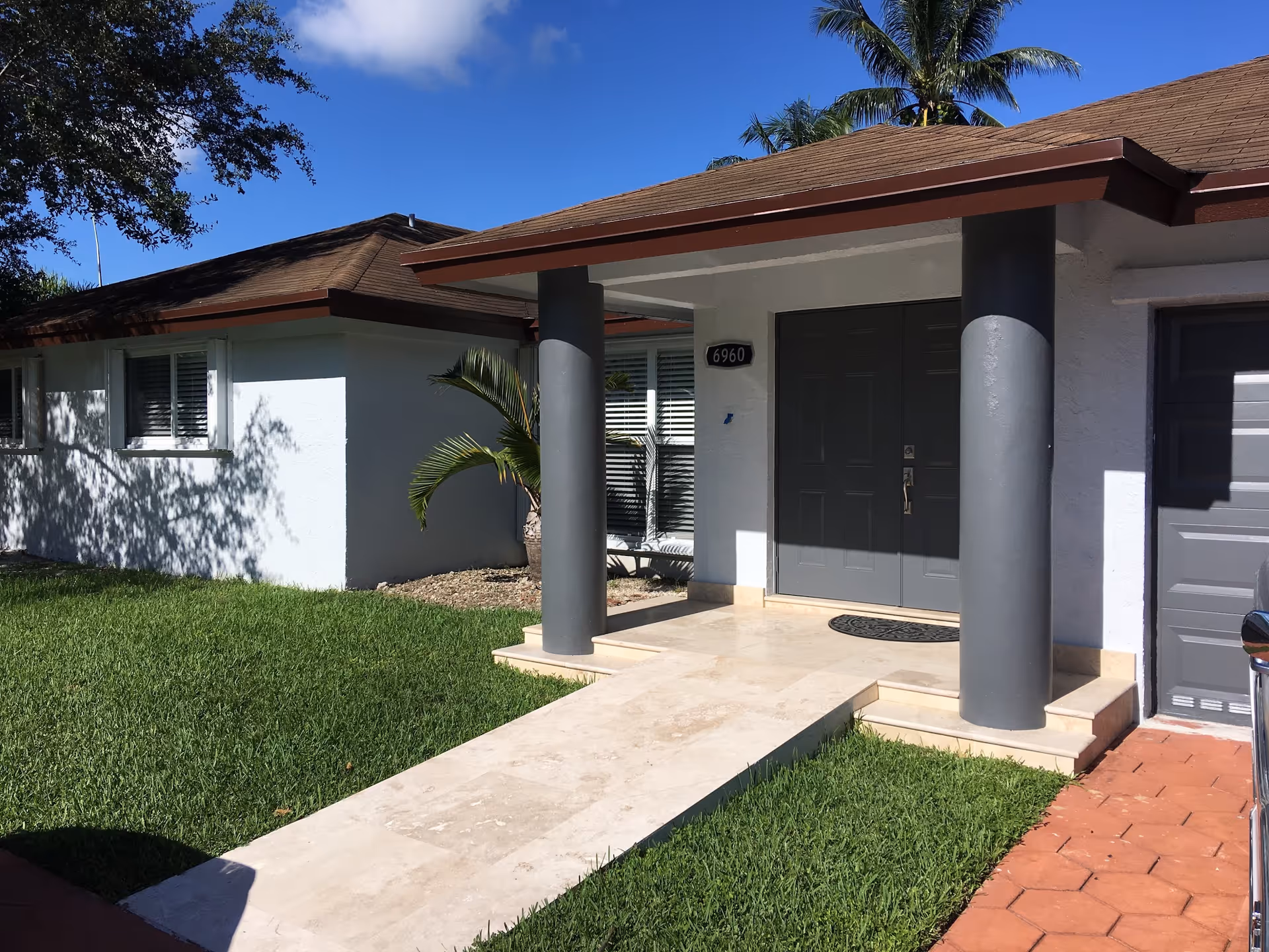 Front entrance of a single-story house with a tiled walkway, two gray columns, double gray doors, and a palm beside a green lawn.