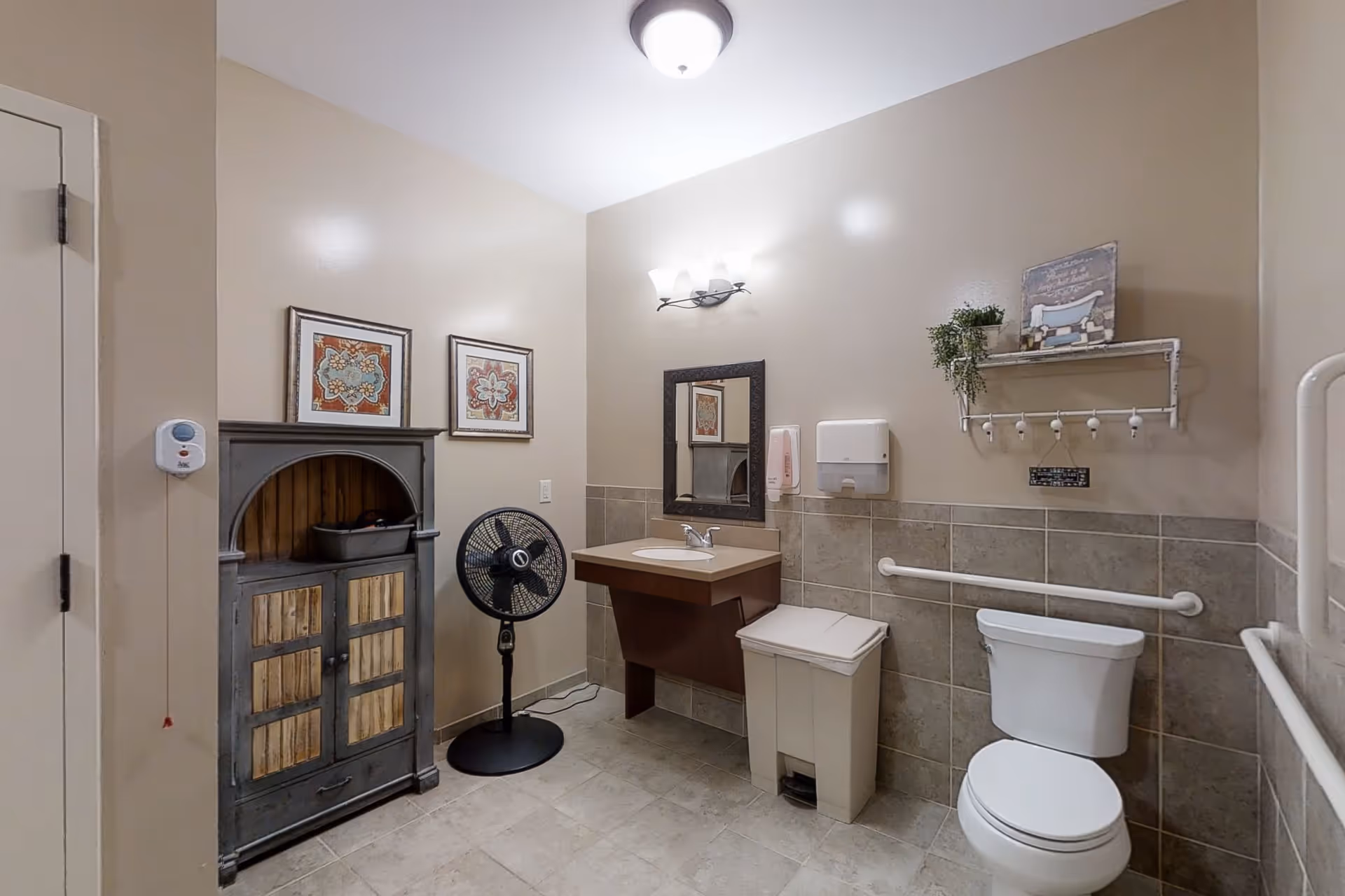 Accessible restroom with a toilet, wall-mounted sink and mirror, grab bars, storage cabinet, shelf and a standing fan.