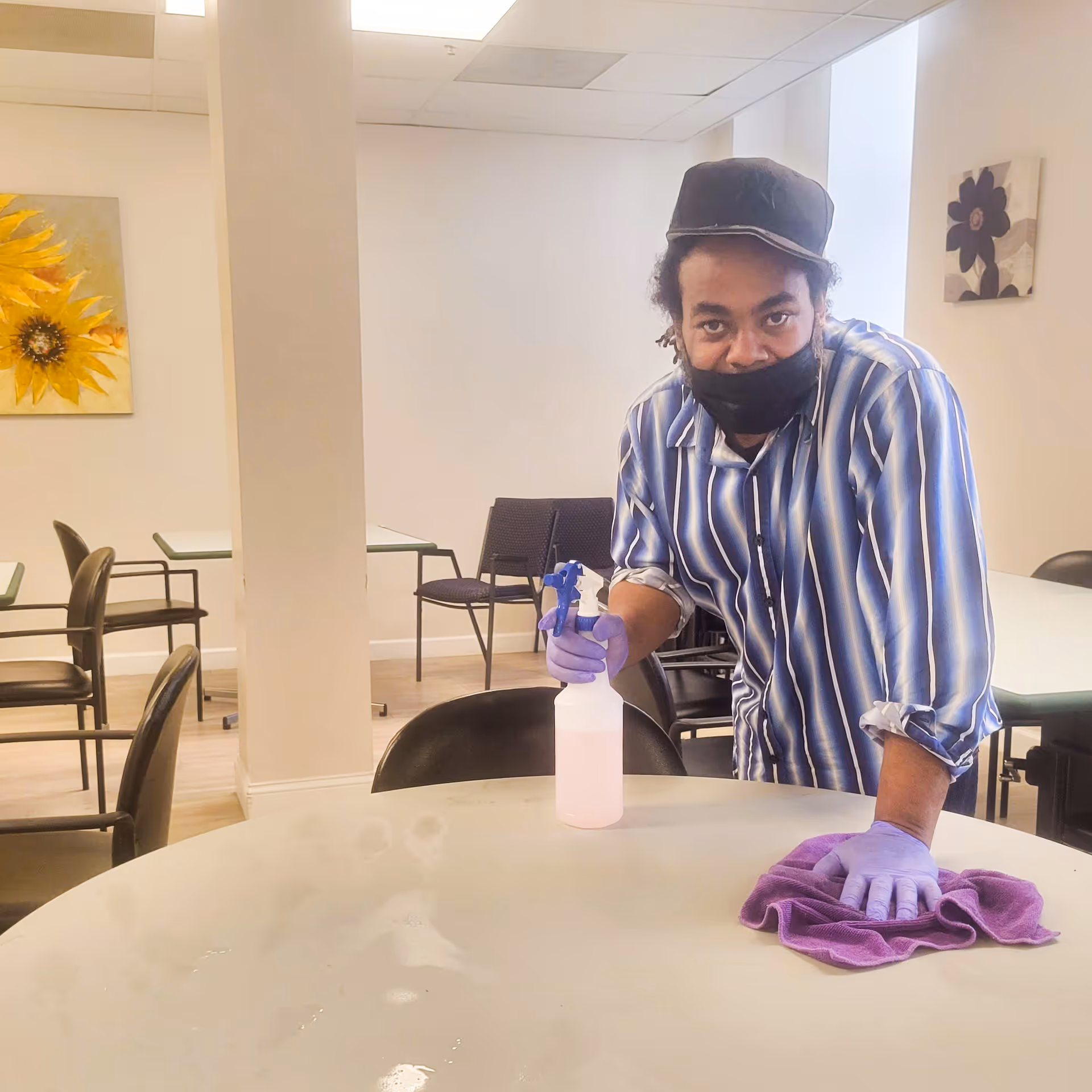 A person wearing a striped shirt, black cap, black face mask, and purple gloves is cleaning a round table with a purple cloth and a spray bottle in a room with multiple tables and chairs. The room has light-colored walls with sunflower and flower paintings.