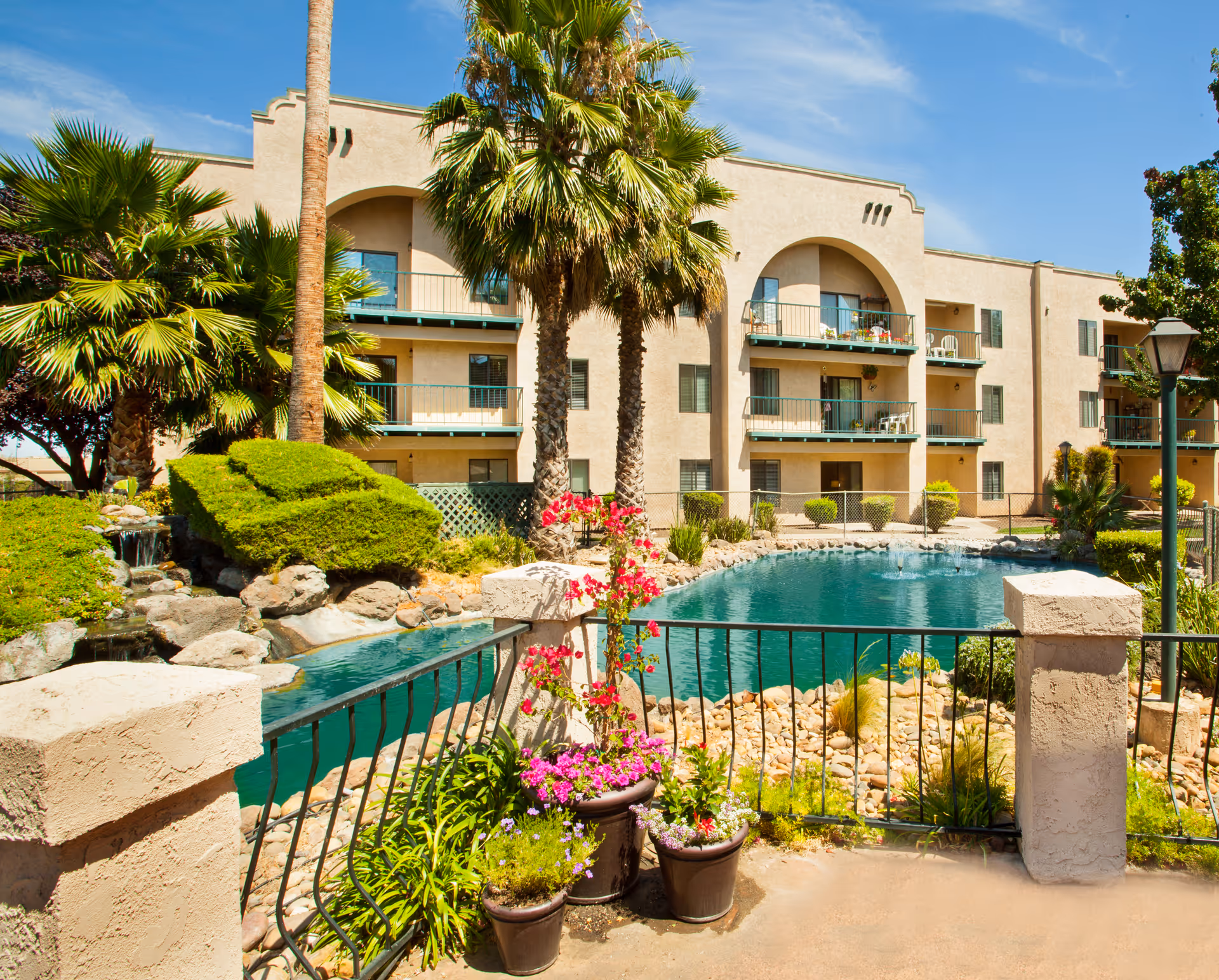 Exterior view of a three-story beige apartment building with balconies overlooking a landscaped pond, palm trees, and potted flowers.