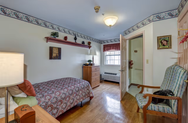 A cozy bedroom in a senior living facility featuring a single bed with patterned bedding, a wooden nightstand with a lamp, a wooden dresser with a lamp and decorative items, a window with a valance, a wooden chair with a plaid cushion, and an open door leading to a bathroom.