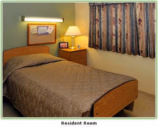 A small resident bedroom with a single bed, bedside table and lamp next to a curtained window.