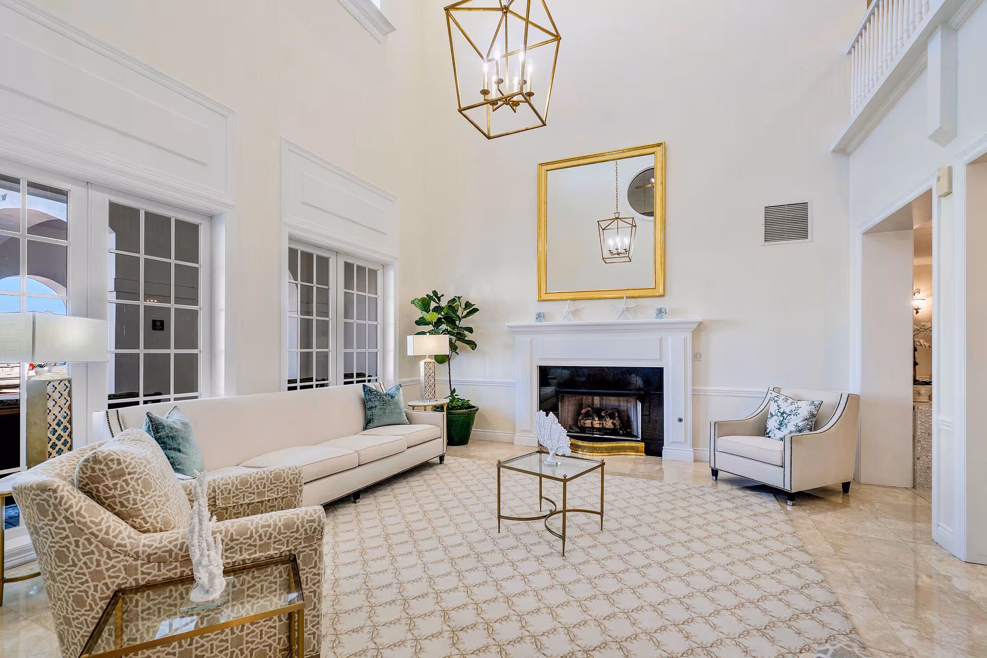 Bright, airy living room with high ceilings, a fireplace, long white sofa, armchairs, patterned rug, and a gold-framed mirror.