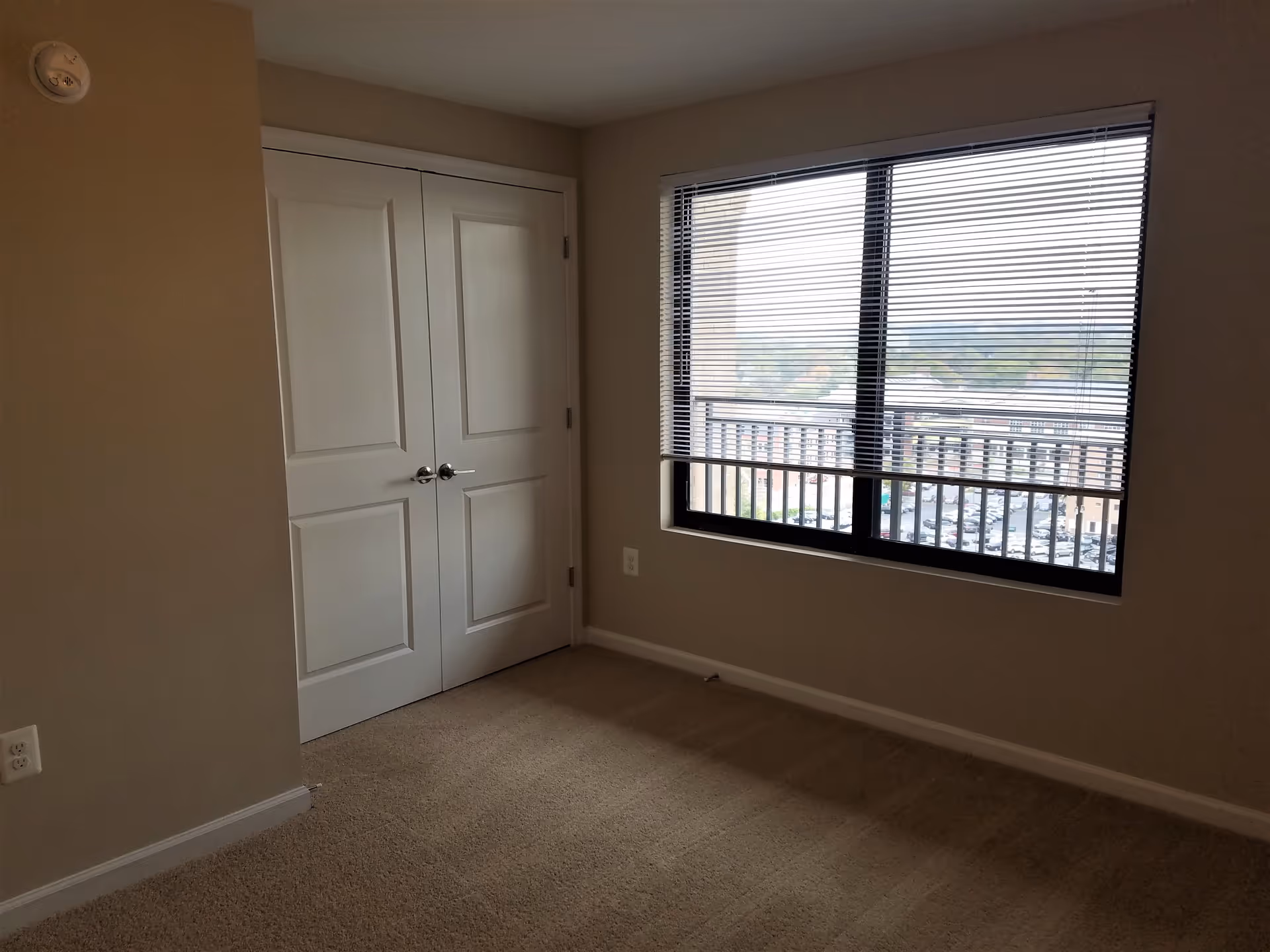 Empty room with beige walls and carpet, a large window with horizontal blinds partially open showing an outdoor view, and a closed double-door closet with white doors and silver handles.