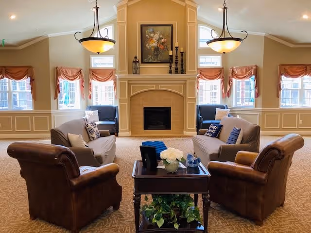 A cozy living room with two brown leather armchairs and two gray sofas arranged around a wooden coffee table with a flower vase and decorative items. The room features a fireplace with a floral painting above it, two hanging light fixtures, and multiple windows with peach-colored valances letting in natural light.