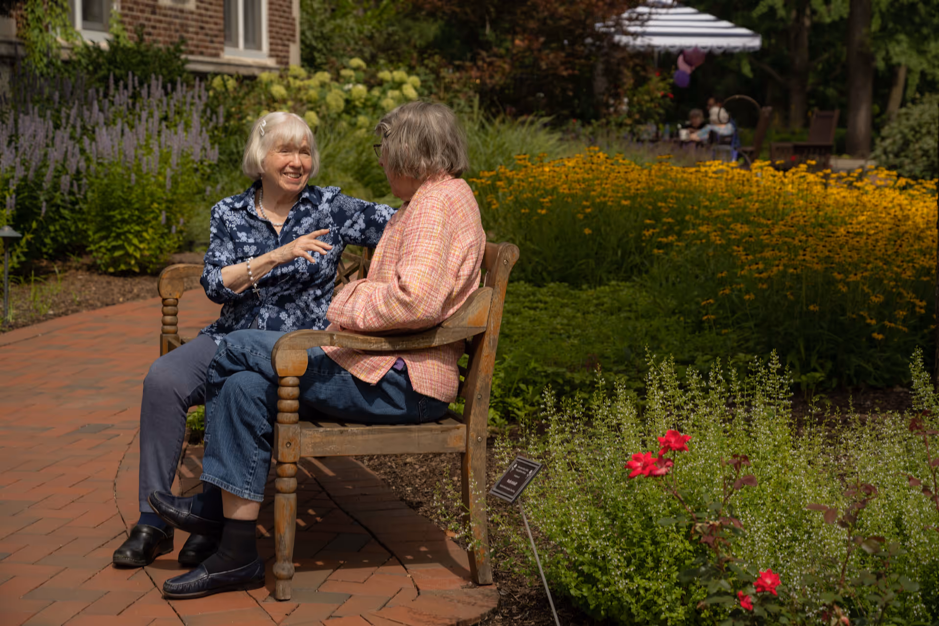 Two elderly women sitting on a wooden bench in a garden area, engaged in conversation. One woman is wearing a blue floral shirt and the other a pink plaid jacket. They are surrounded by green plants and colorful flowers, with a brick pathway beside them.