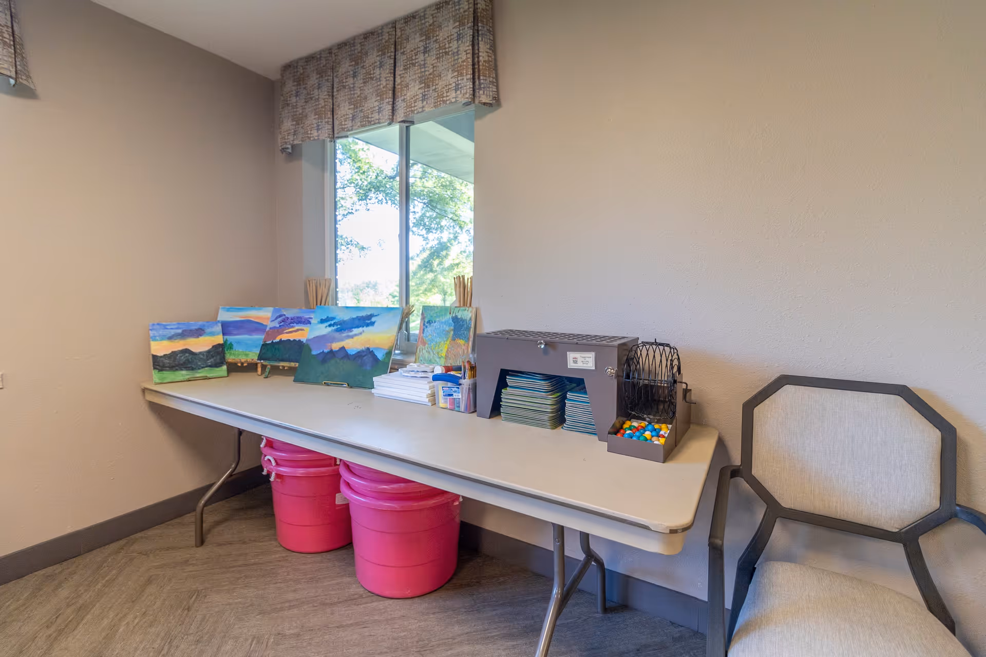 A small activity room with a folding table against the wall holding several landscape paintings, art supplies, and a bingo cage with colorful balls. Two large pink buckets are under the table. A beige cushioned chair with dark wooden arms is placed next to the table. A window with a patterned valance lets in natural light.