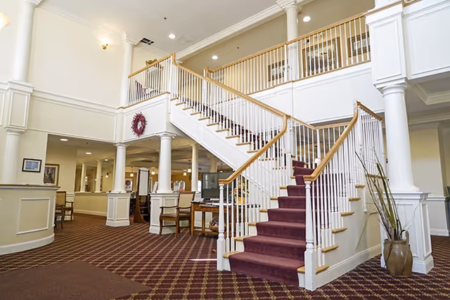Interior view of a senior living facility lobby featuring a grand staircase with red carpet, white railings, and wooden handrails. The area has beige walls, white columns, patterned carpet flooring, and several chairs and tables arranged around the space. A decorative wreath hangs on the wall above the staircase.