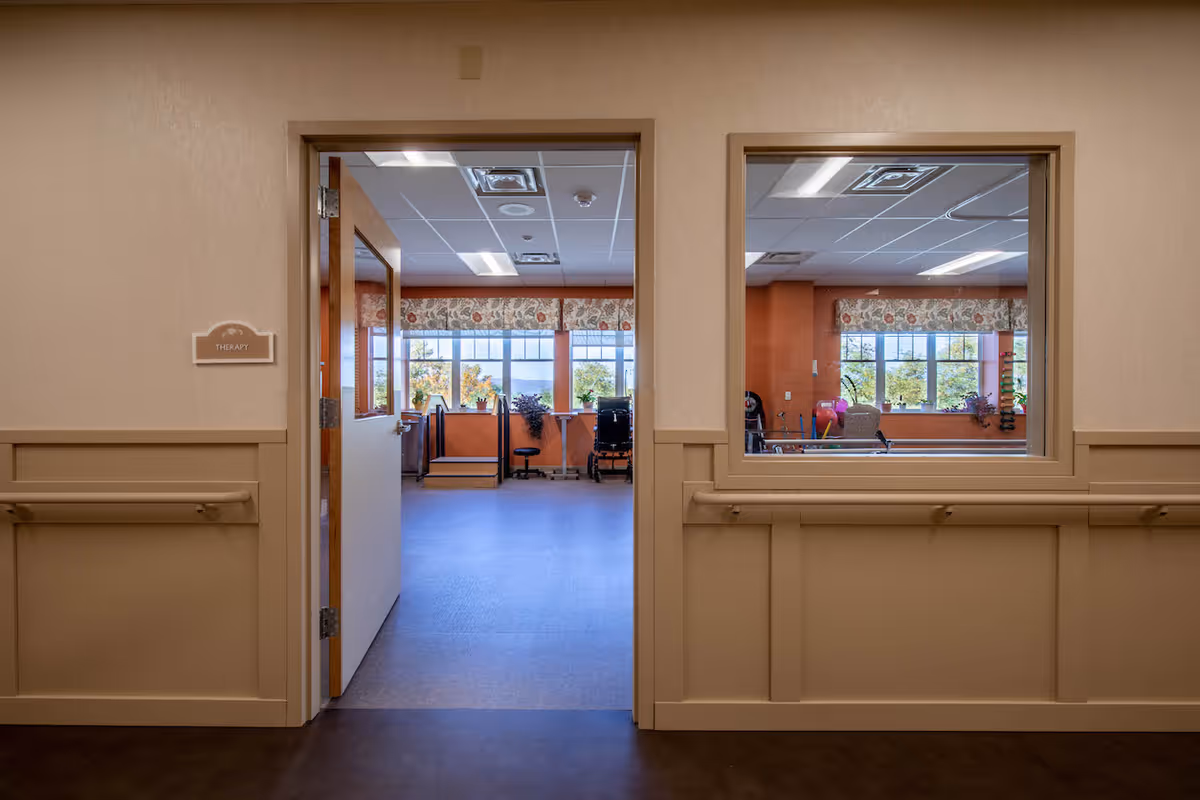 View through an open door and window into a therapy room with exercise equipment and large windows with floral valances, seen from a hallway with beige walls and handrails.