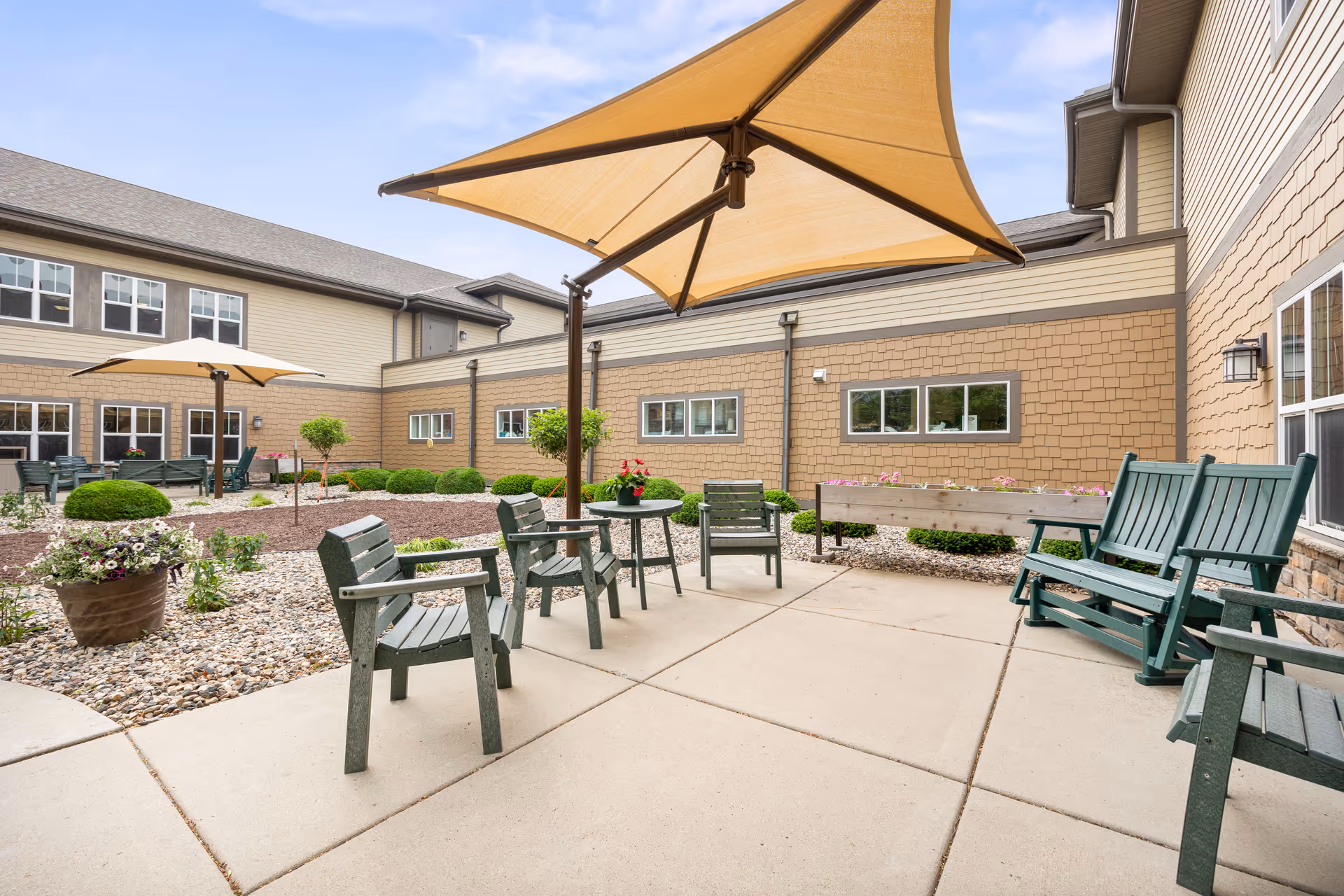 Outdoor courtyard patio with green wooden chairs, large beige umbrellas, potted plants, and a surrounding two-story building.