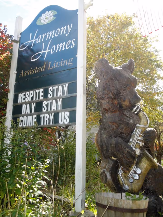Outdoor sign for Harmony Homes Assisted Living with a message board reading 'Respite Stay Day Stay Come Try Us' next to a wooden bear sculpture playing a saxophone, surrounded by greenery and trees.