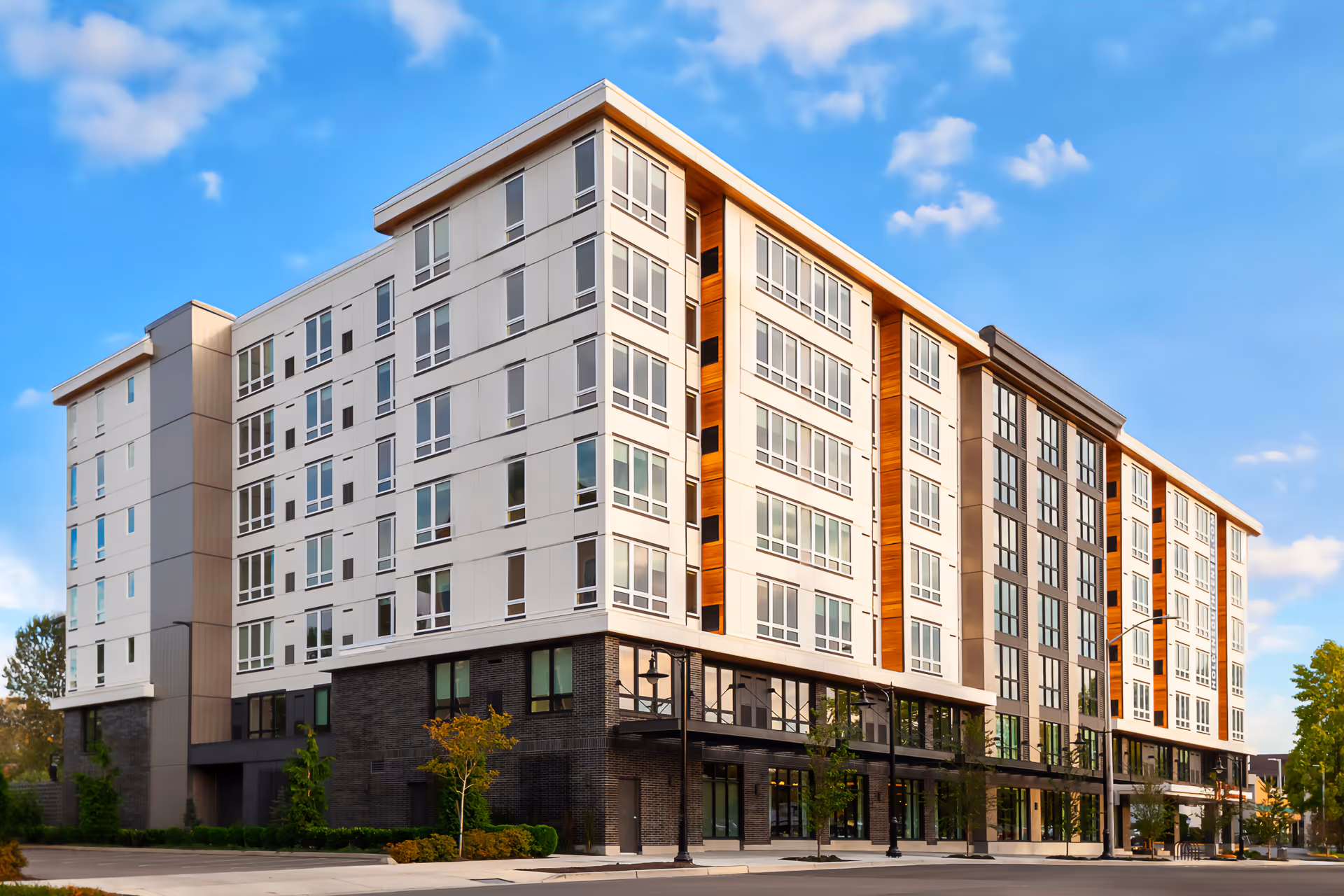 Exterior view of a modern multi-story senior living facility building with large windows, a mix of white and dark paneling, and some wooden accents under a blue sky with scattered clouds.
