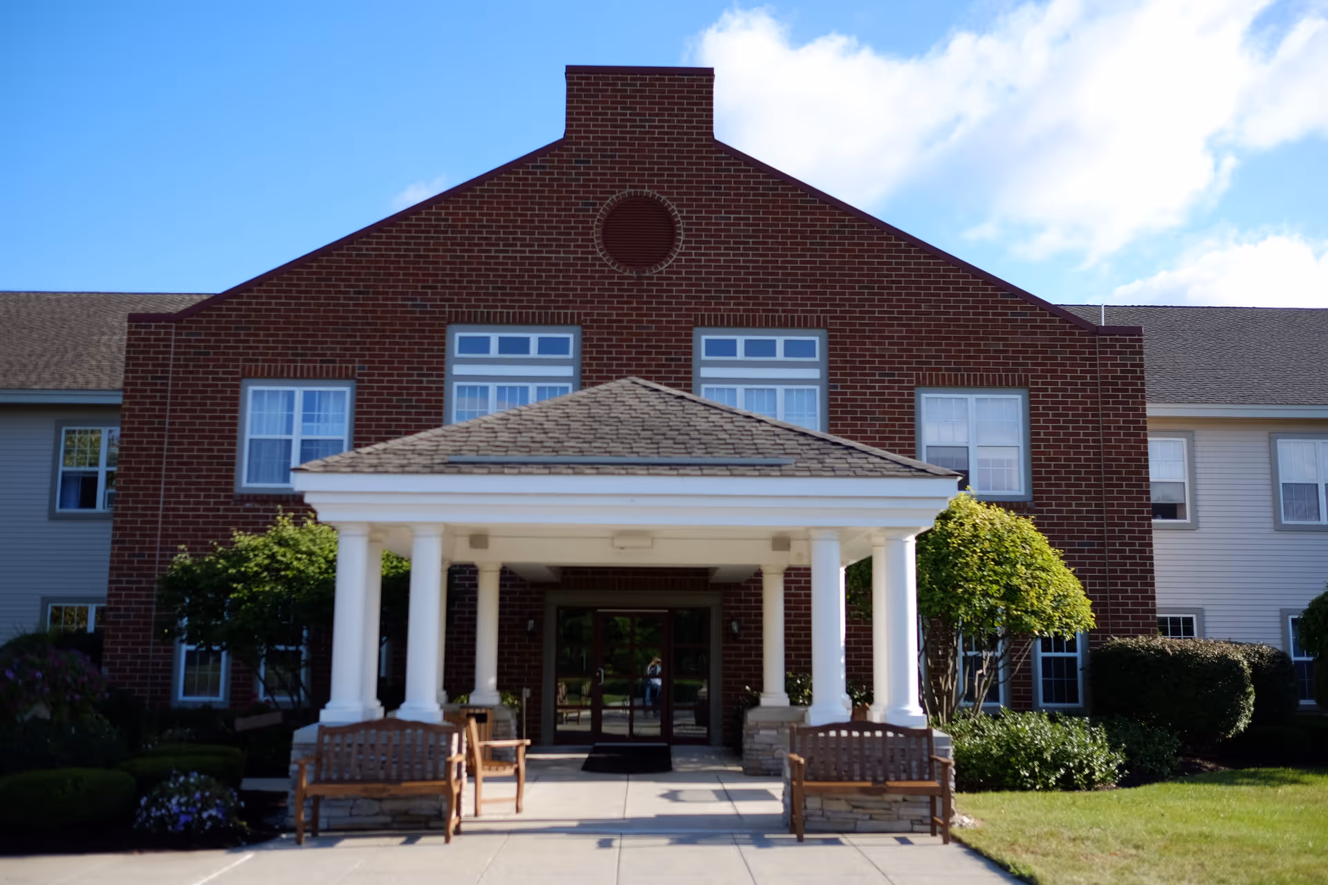 Front exterior view of a senior living facility building with a brick facade, white columns supporting a covered entrance, benches on either side of the walkway, and well-maintained landscaping including bushes and trees under a blue sky with some clouds.
