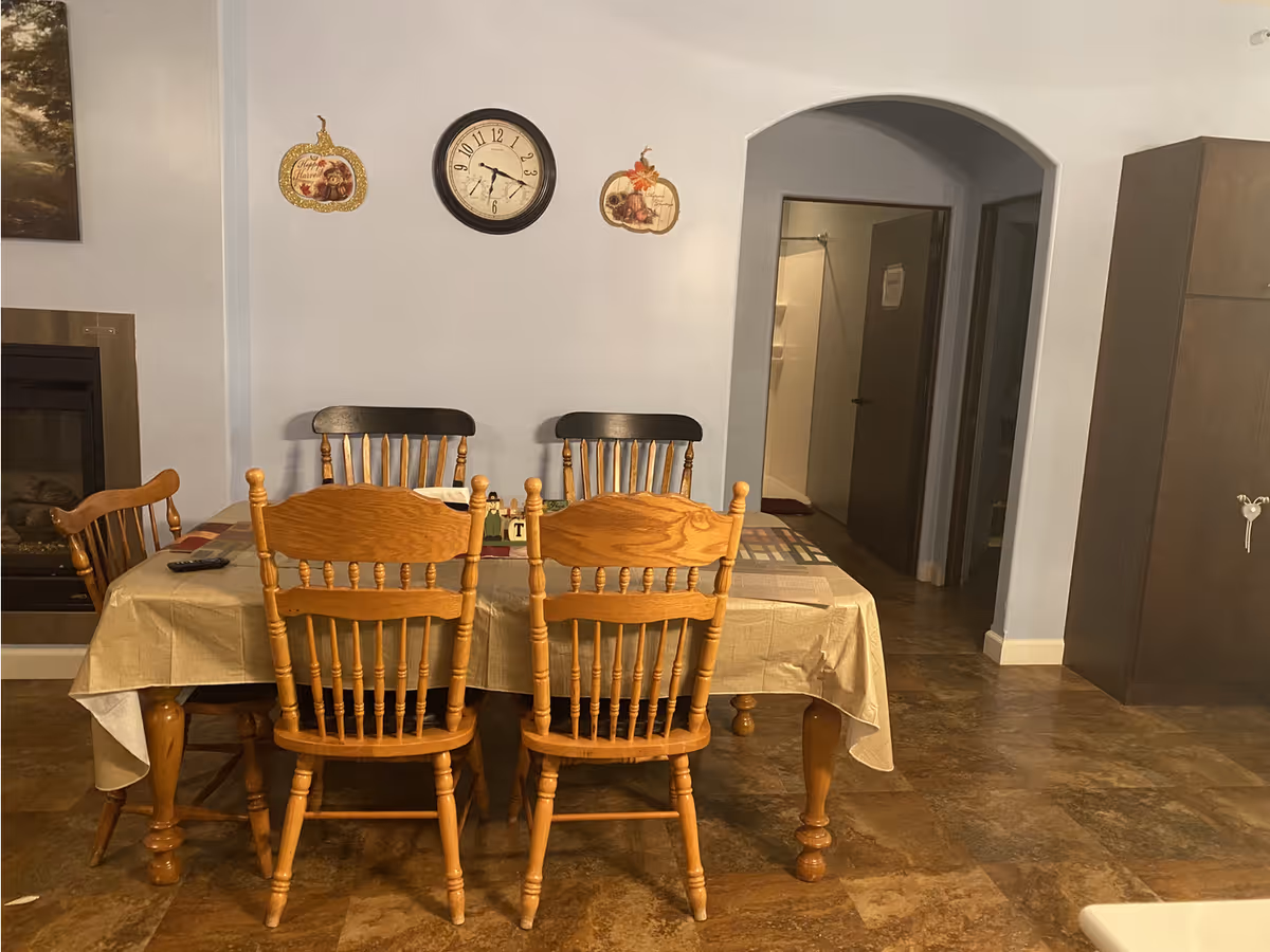A dining area with a rectangular wooden table covered with a beige tablecloth and six wooden chairs around it. On the wall above the table, there is a round clock and two pumpkin-shaped wall decorations. To the left, there is a fireplace, and to the right, there is a tall cabinet. An arched doorway leads to a hallway with open doors.
