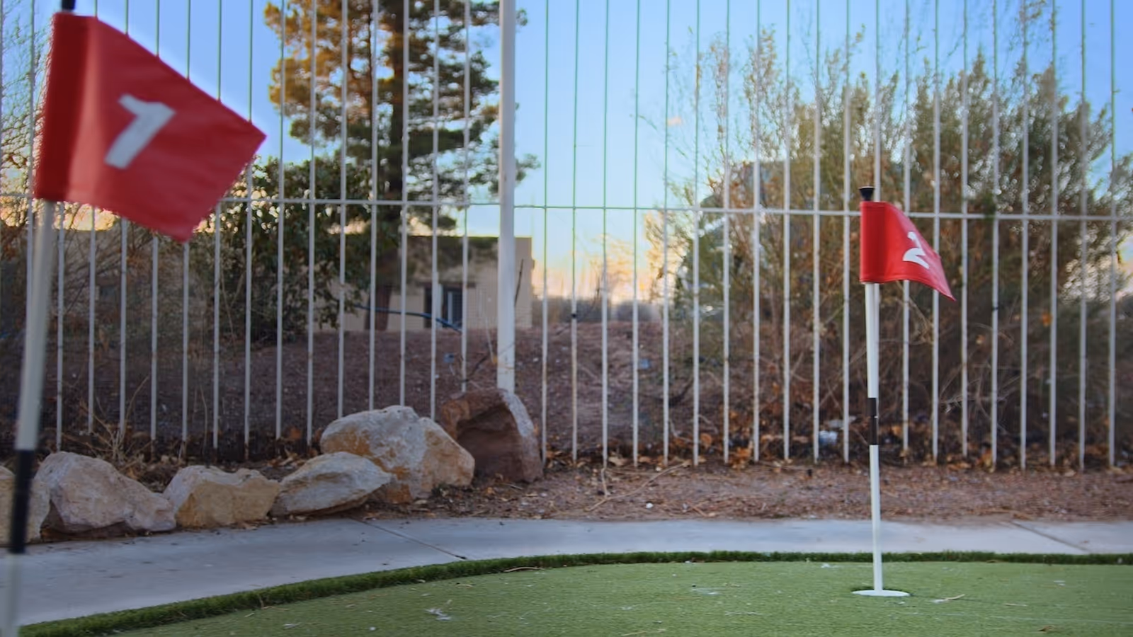 Outdoor mini golf putting green with two red flags numbered 1 and 2, surrounded by rocks and a metal fence with trees and bushes in the background.
