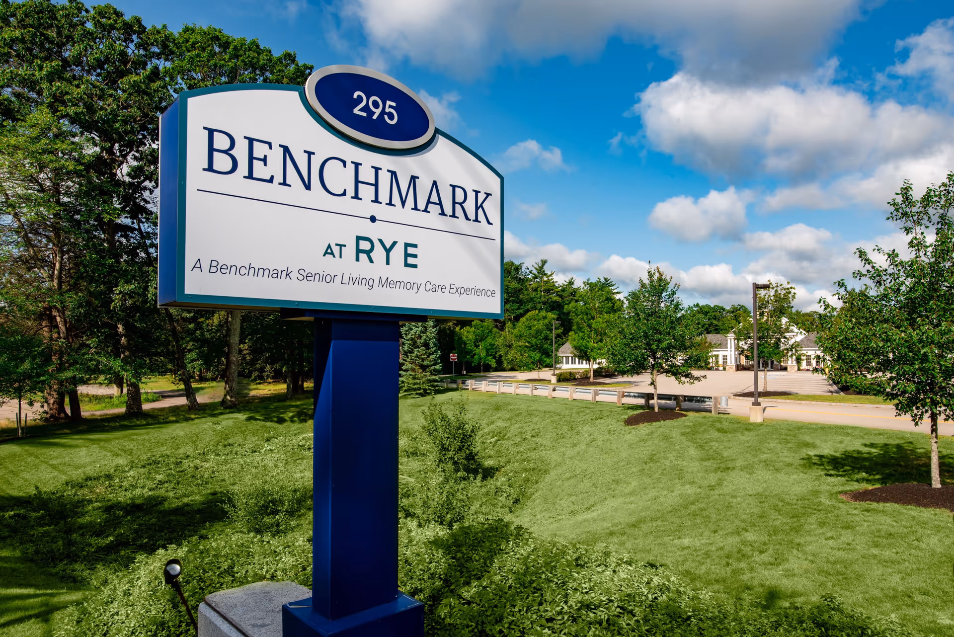 Outdoor view of the Benchmark at Rye senior living facility sign with the address number 295. The sign is mounted on a blue post and surrounded by green grass and trees under a partly cloudy blue sky. In the background, there is a parking lot and part of the facility building.