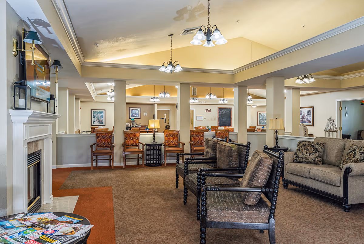 A spacious senior living facility common area with comfortable seating including armchairs and a sofa, a fireplace on the left, and a dining area in the background with chairs and tables. The room features warm lighting from ceiling fixtures and lamps, with framed artwork on the walls.