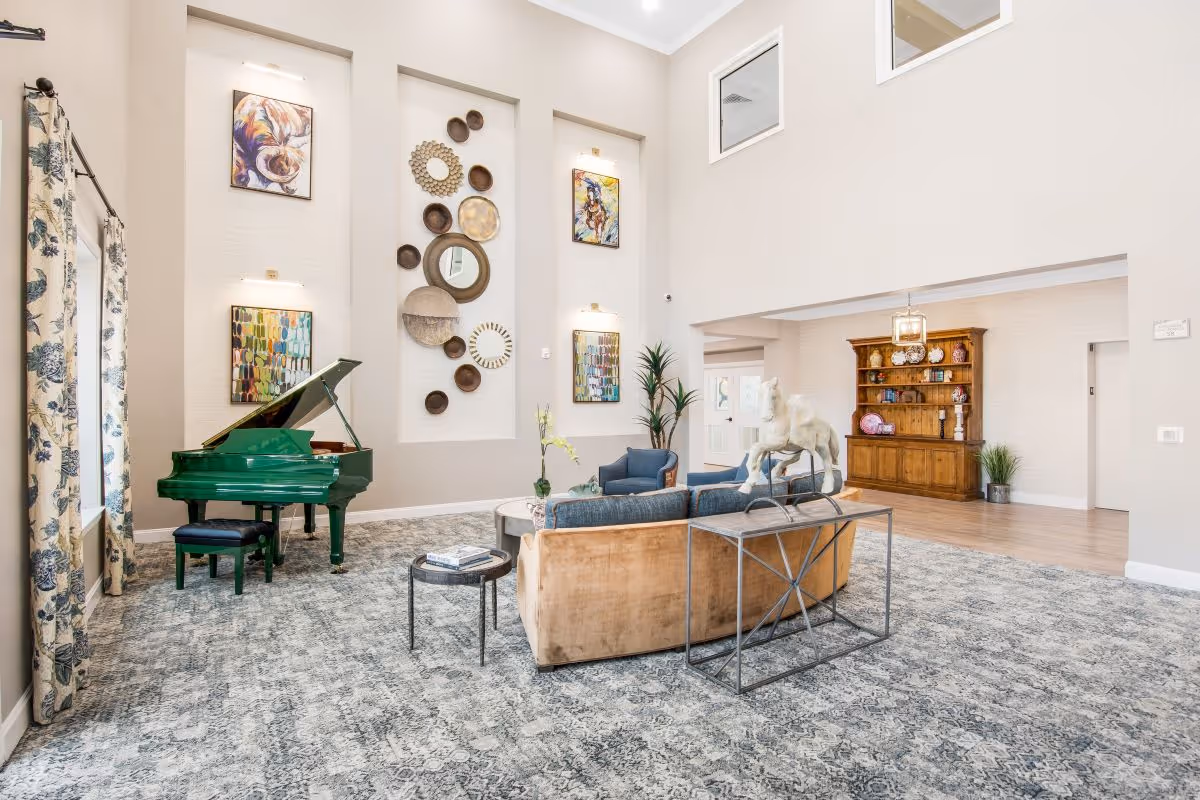 A spacious and well-lit living room area with a green grand piano on the left, a beige sofa with blue cushions, a small round side table, and a decorative console table with a white horse sculpture. The walls feature artistic wall decor and paintings, with tall windows and floral curtains on the left side. In the background, there is a wooden cabinet with decorative items and a hanging light fixture.