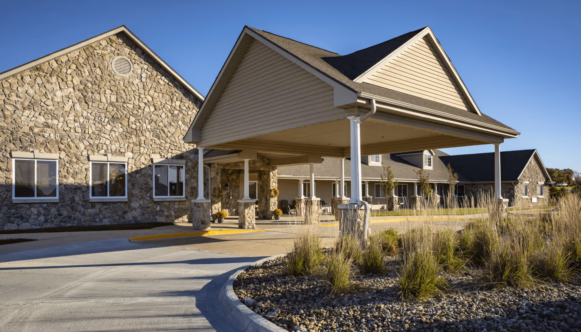 Exterior view of a senior living facility with stone and siding walls, a covered entrance supported by columns, and a landscaped area with tall grasses and rocks under a clear blue sky.