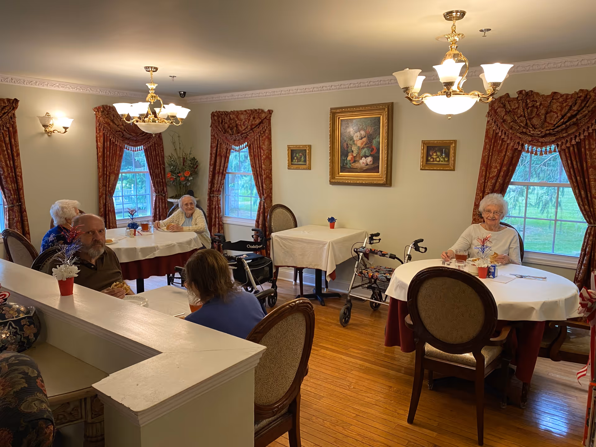 A dining room in Colonial Manor Hackettstown with elderly residents seated at round tables covered with white tablecloths. The room has wooden floors, large windows with red patterned curtains, and framed paintings on the walls. There are walkers near some tables and decorative light fixtures hanging from the ceiling.