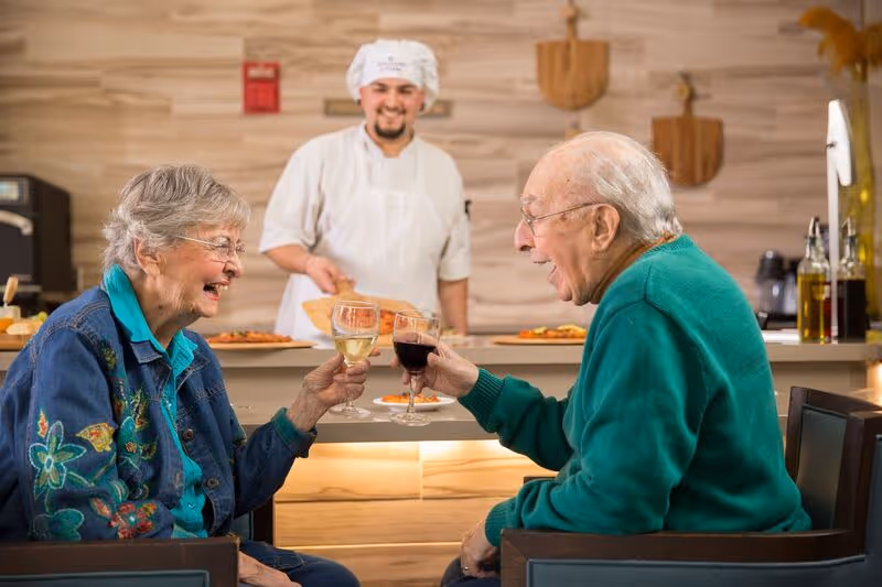 An elderly woman and an elderly man sitting at a table in a dining area, smiling and clinking wine glasses. A chef in a white uniform and hat stands behind the counter holding a pizza, smiling at them.