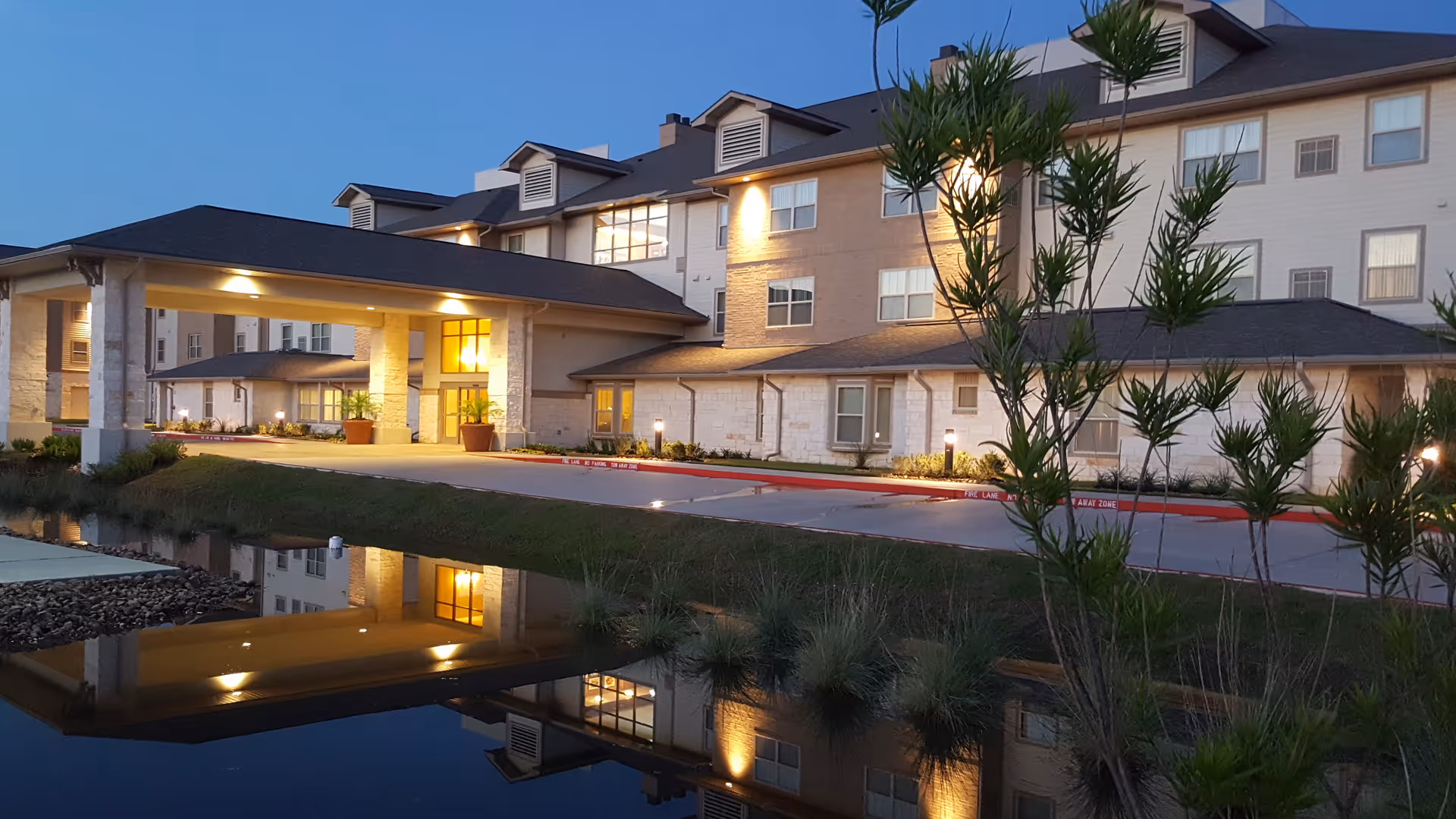 Exterior view of Oak Park Retirement Resort building at dusk with lights on, showing a covered entrance and a reflecting pond in the foreground.