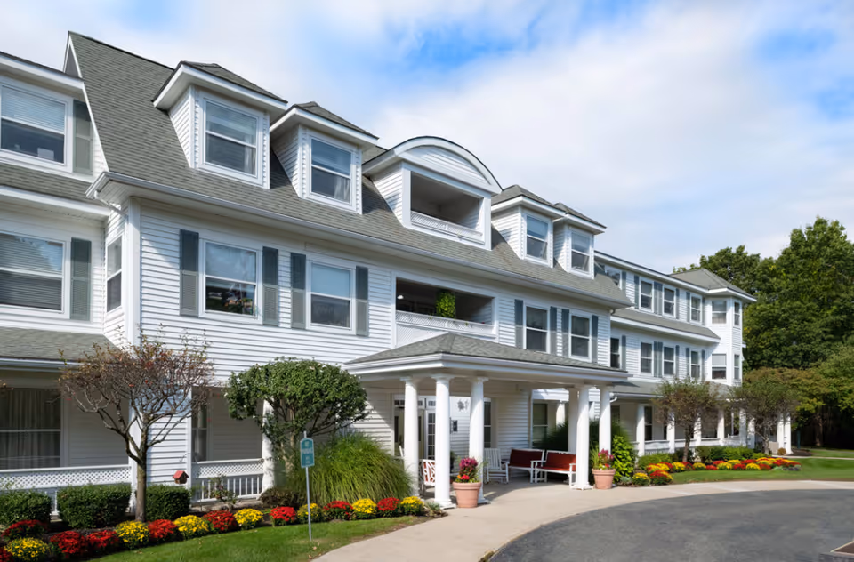 Exterior view of a large, white senior living facility building with multiple windows and a covered entrance supported by white columns. The building is surrounded by well-maintained landscaping including bushes, small trees, and colorful flowers. The sky is partly cloudy.