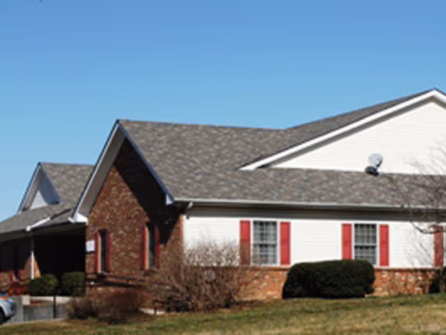 Single-story brick-and-siding assisted living building with red shutters, shrubs, and a clear blue sky.
