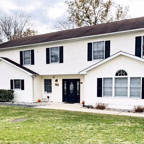 Exterior view of a two-story white residential building with black shutters and a dark roof. The building has a central entrance with double doors and a small porch area. There is a grassy lawn in front and some small shrubs near the building. Trees with bare branches are visible in the background under a cloudy sky.