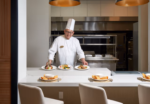 A chef wearing a white uniform and tall white hat is placing three plates of food on a counter in a kitchen setting. The counter has place settings with plates, bowls, and cutlery, and there are three chairs in front of the counter. The background shows stainless steel kitchen appliances and equipment.