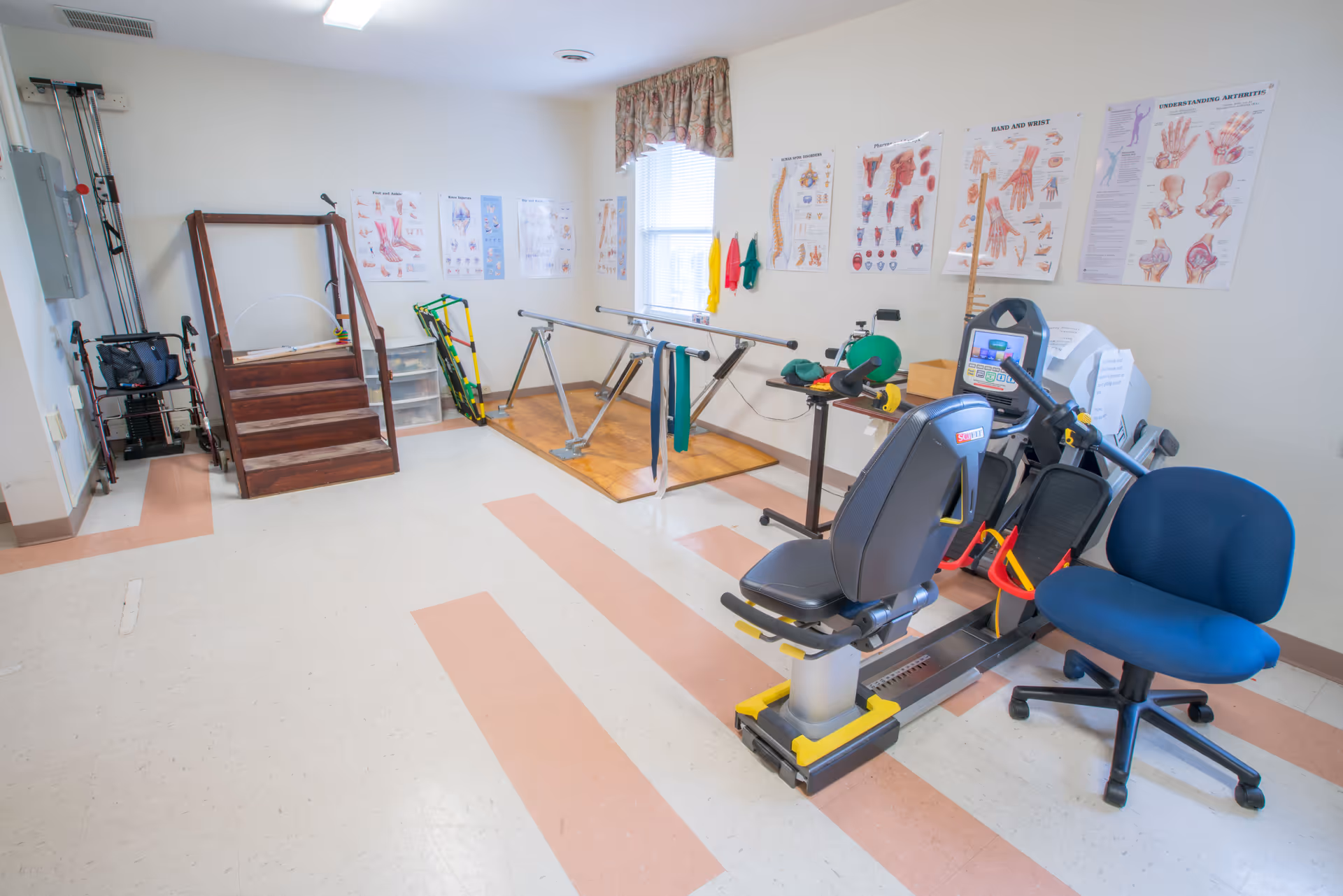 A physical therapy room in a nursing center with exercise equipment including a recumbent bike, parallel bars, a small wooden staircase for step exercises, and various therapy tools. Anatomical charts are displayed on the walls, and a blue office chair is positioned near the exercise machines.