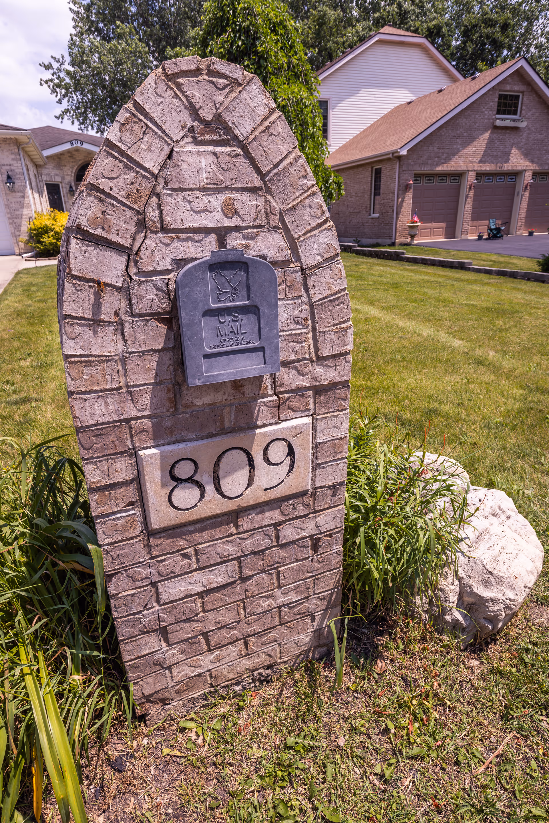 A brick mailbox structure with the house number 809 displayed on a stone plaque. The mailbox is mounted on the brick structure, and there is grass and plants surrounding the base. In the background, there are residential houses with garages and well-maintained lawns under a partly cloudy sky.