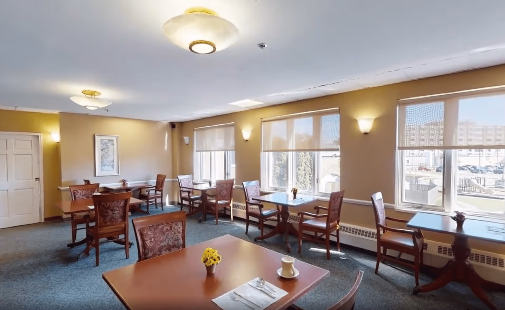 A bright dining room with several wooden tables and chairs arranged neatly. Each table has a small flower vase, and one table has a cup and napkin. Large windows with roller shades allow natural light to fill the room. The walls are painted beige with wall-mounted light fixtures and a framed picture on one wall.