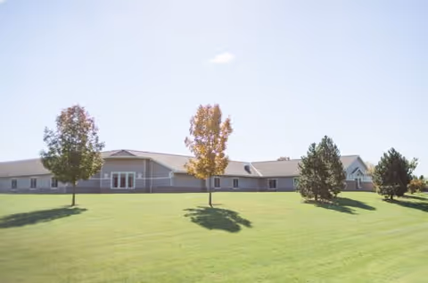 A single-story building with a light-colored exterior situated on a large, well-maintained grassy lawn with a few trees scattered around. The sky is clear with minimal clouds.