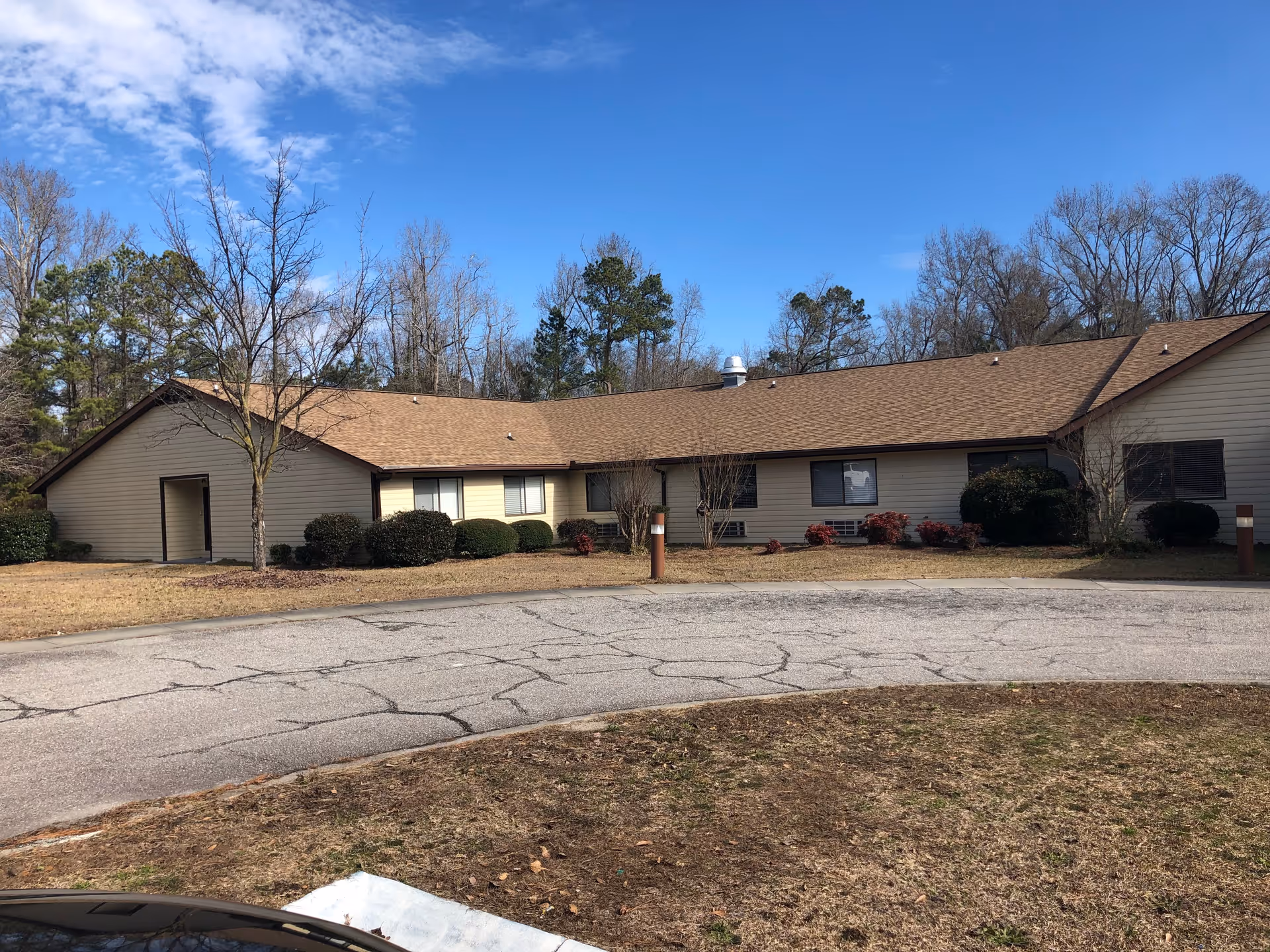 Single-story beige building with a brown roof, shrubs and a circular cracked driveway under a blue sky.