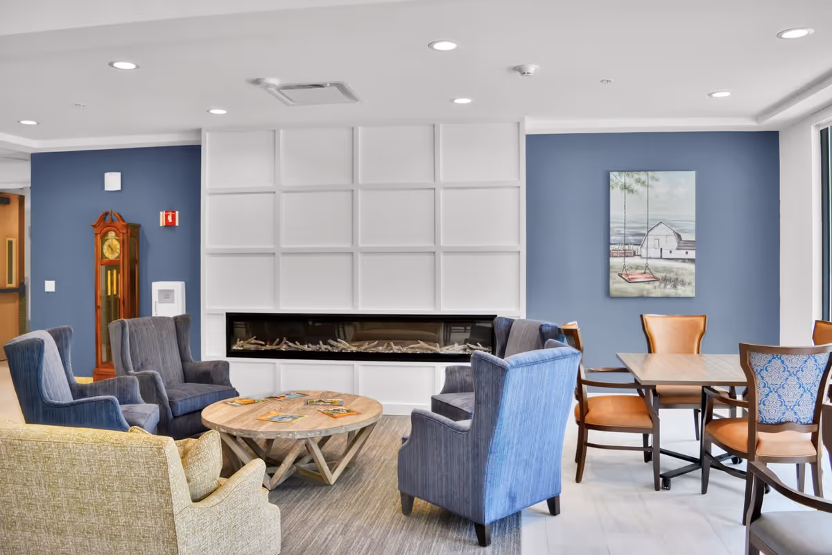 A bright common lounge with armchairs arranged around a round coffee table and a linear fireplace set into a white paneled wall.