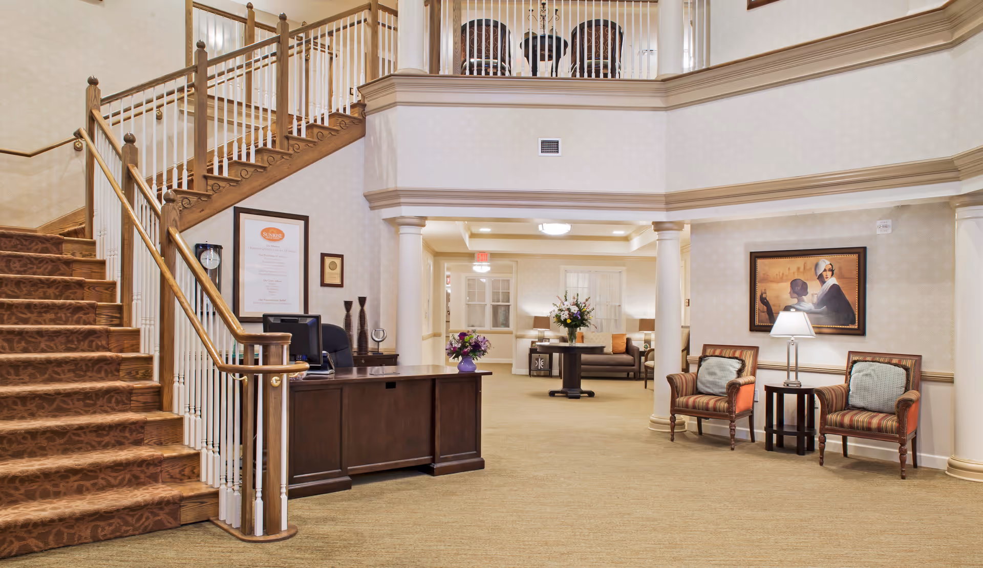 Spacious senior living lobby with a grand staircase, reception desk, seating area and decorative columns.